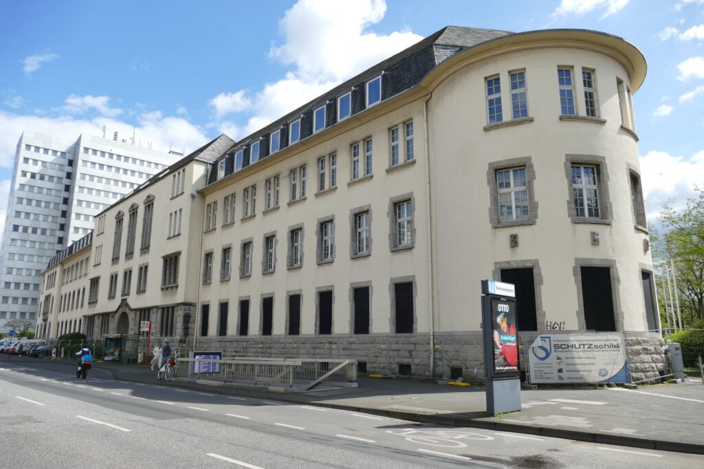 cremefarbenes, dreistöckiges, altes Gebäude mit turmartig abgerundeter Ecke; davor ein Haltestellenschild, ein Treppenabgang und eine Bushaltstelle. Im Hintergrund ein funktionales Hochhaus.